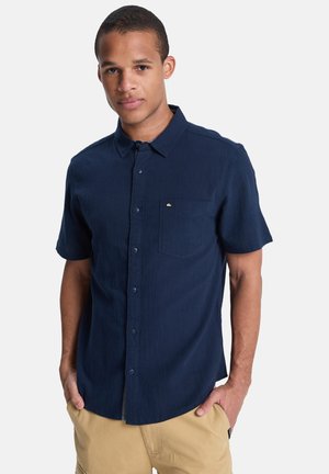 Camicetta blu navy a maniche corte realizzata in tessuto testurizzato, con chiusura a bottoni e tasca sul petto, caratterizzata da un piccolo logo.