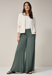 Cream zip-up bomber jacket over a white shirt, paired with wide-leg, olive-green pants. Model wears black slide sandals. Smooth fabric textures.