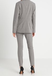 Šedý kostkovaný oblek s přiléhavým sakem a zúženými kalhotami. Sako má klopový límec a outfit doplňují černé vysoké boty na podpatku.