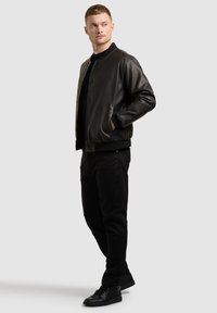 Junger Mann, der eine schwarze Leder-Bomberjacke, schwarze Hose und schwarze Turnschuhe trägt, steht mit einer Hand in der Tasche und schaut seitlich.