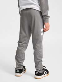 Kind draagt een grijze trainingsbroek met witte chevron-logo's aan de zijkant, zwart-witte sneakers, staand met de rug naar de camera.