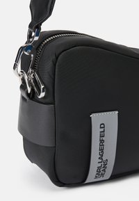 Črn najlon crossbody nahrbtnik z gladko teksturo, srebrno kovinsko opremo, zadrgo in sivo oznako z logotipom "KARL LAGERFELD JEANS."
