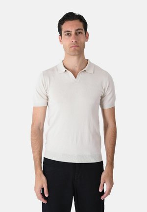 Uomo con capelli corti e scuri che indossa una polo beige chiaro a maniche corte e pantaloni neri, in piedi davanti a uno sfondo bianco uniforme.