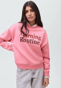 Žena s dlouhými tmavými vlasy, oblečená v růžové mikině s nápisem "Ranní rutina" a světlých šedých džínách, stojí s rukou na bocích.