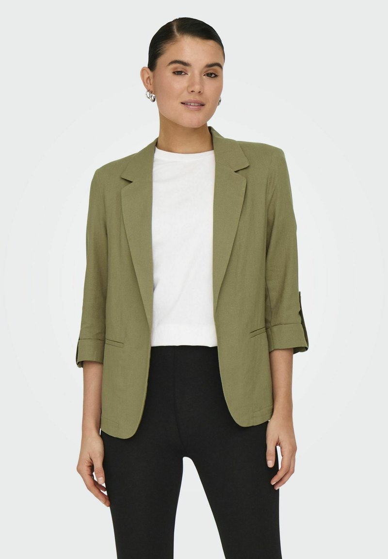 Blazer in lino verde oliva con una vestibilità su misura, revers classici e maniche a 3/4, abbinato a una t-shirt bianca e pantaloni neri.