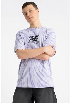 Młody mężczyzna w jasnofioletowej koszulce tie-dye z czarnym napisem i grafiką, z założonymi rękami, stoi na białym tle.