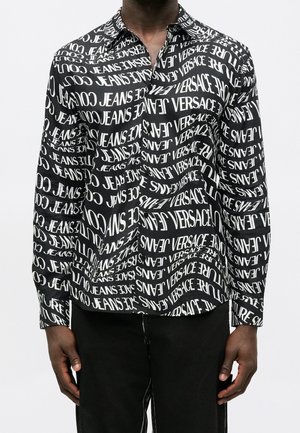 Czarna koszula na długi rękaw z guzikami, z białymi powtarzającymi się wzorami tekstu z napisem "VERSACE JEANS COUTURE", noszona przez osobę w czarnych spodniach.