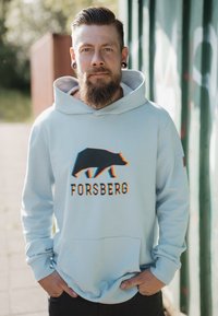 Felpa blu chiaro con un grafico di un orso nero e il testo "FORSBERG". Presenta una tasca frontale e un cappuccio con lacci. Tessuto morbido.