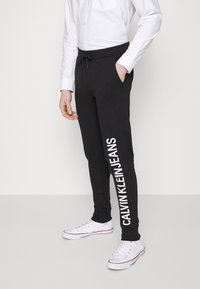 Černé tepláky s nastavitelným pasem a bílým nápisem "CALVIN KLEIN JEANS" podél levé nohy. Nosené s bílými teniskami.