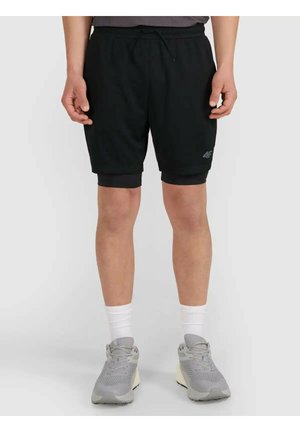 Osoba nosząca czarne, warstwowe sportowe spodenki, białe skarpetki za kostkę oraz szare buty do biegania, stojąca na jednolitym tle.