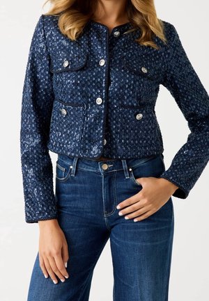 Vrouw draagt een marineblauw paillettenjasje met knopen en vier zakken en donkerblauwe spijkerbroek, één hand op de heup, tegen een witte achtergrond.