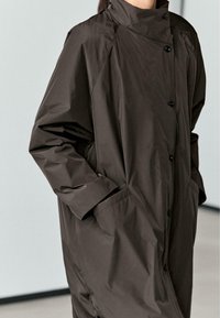 Brauner Oversize-Mantel mit hohem Kragen, ausgestattet mit einer Knopfleiste und Seitentaschen. Hergestellt aus einem glatten, wasserabweisenden Stoff.