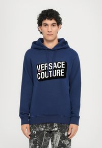 Felpa con cappuccio color blu navy realizzata in cotone misto, con un grande logo grafico a contrasto in bianco e nero sulla parte anteriore. Modello che indossa pantaloni con fantasia.