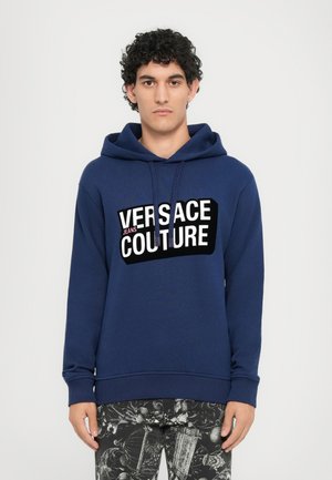 Navy blue hoodie made of cotton blend with a large contrasting black and white graphic logo on the front. Model wearing patterned pants.
