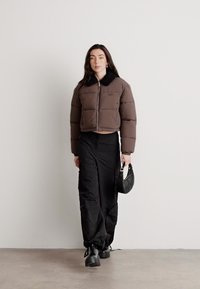 Brązowa, krótka kurtka puchowa z czarnym kołnierzem, zestawiona z czarnymi spodniami cargo i małą torebką. Modelka nosi czarne botki. Gładkie struktury.
