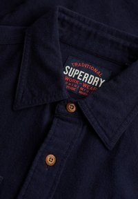 Chemise bleu foncé à col avec des boutons en bois visibles et une étiquette Superdry Workwear cousue à l'intérieur du col.