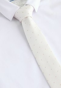 Camisa blanca de vestir con una corbata de color beige claro texturizada que presenta pequeños lunares uniformemente espaciados, anudada con un nudo clásico.