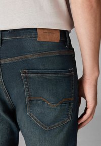 Tamno plave traperice s leđnom džepom s kontrastnim narančastim šavom i smešnom kožnom etiketom koja glasi "DENIM SUPPLY Co."
