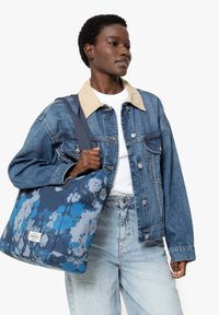 Veste en denim avec un col beige, un t-shirt blanc et un jean bleu clair. Portant un sac fourre-tout tie-dye bleu avec des accessoires à l'avant.