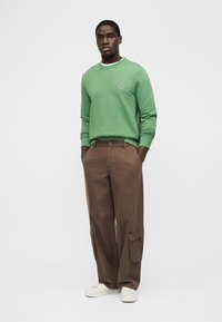 Jeune homme debout, les mains dans les poches, portant un sweat-shirt vert, un pantalon cargo marron et des baskets blanches, sur un fond uni.
