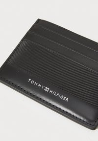 Tarjetero de piel negra con un diseño de rayas diagonales texturizadas, múltiples compartimentos para tarjetas y el logo en relieve "TOMMY HILFIGER".