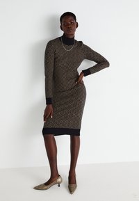 Vestido de malha com um padrão geométrico em preto e dourado, com um decote alto e punhos canelados. Combinado com sapatos de salto agudo dourados.