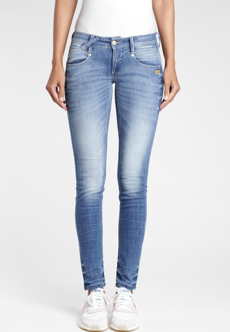 Jeans skinny en denim bleu clair, avec un design décoloré, des poches avant et une fermeture éclair avec un bouton. Portés avec des baskets blanches.