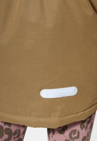 Jupe en tissu marron avec une apparence texturée, comportant un patch logo Hummel blanc à l'ourlet. En dessous, des leggings imprimés léopard rose sont visibles.