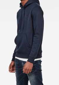 Felpa blu navy realizzata in tessuto morbido, con tasca a marsupio, cappuccio con cordino e polsini a coste. Indossata sopra una maglietta bianca.