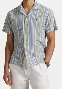 Košile na knoflíky s krátkým rukávem, světle modrá se zelenými, červenými a bílými pruhy; má kempový límec a malou nášivku na levé straně hrudníku.