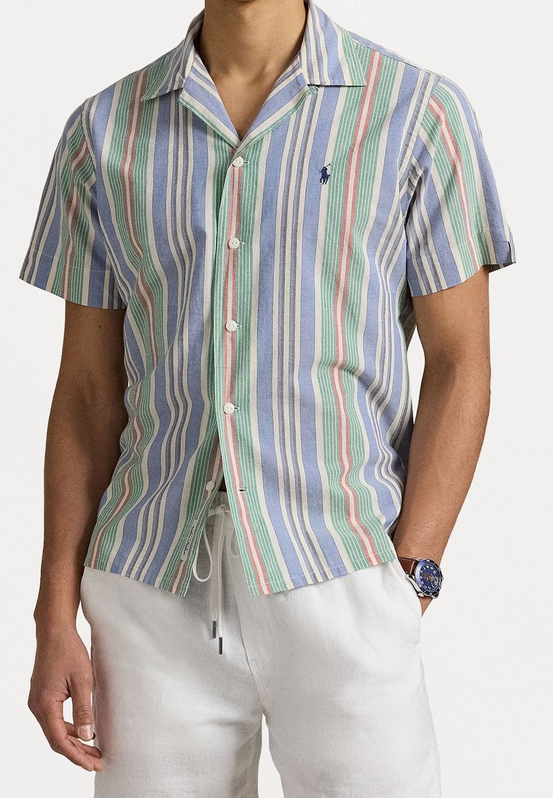 Košile na knoflíky s krátkým rukávem, světle modrá se zelenými, červenými a bílými pruhy; má kempový límec a malou nášivku na levé straně hrudníku.