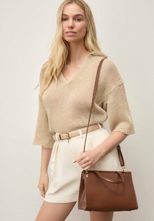 Blonde vrouw in beige gebreide top en crèmekleurige rok met een bruine leren handtas met schouderriem tegen een effen achtergrond.