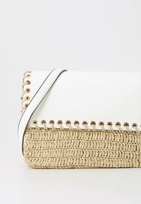 Pleteni slamni torba s bijelim kožnim preklopom, ukrašena zlatnim metalnim detaljima i obrubom s metalnim ojačanjima. Uključen je podesivi remen.