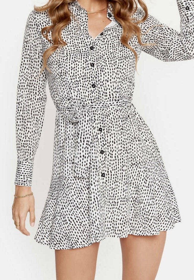 Vestito bianco a maniche lunghe con bottoni, caratterizzato da un motivo a pois neri irregolari, con vita cintata e gonna svasata, indossato da una donna con capelli mossi.