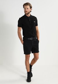 Polo negro con cuello y tres botones, combinado con pantalones cortos negros. El modelo lleva un cinturón negro y zapatillas negras texturizadas.