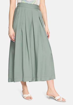 Femme portant une jupe midi plissée vert sauge clair associée à des sandales blanches à talons ouvertes, debout devant un fond uni.