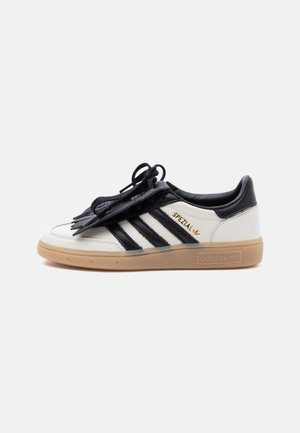 Baskets Adidas Spezial blanches et noires avec semelle en gomme, bandes noires et cache-lacets noir sur un fond clair uni.