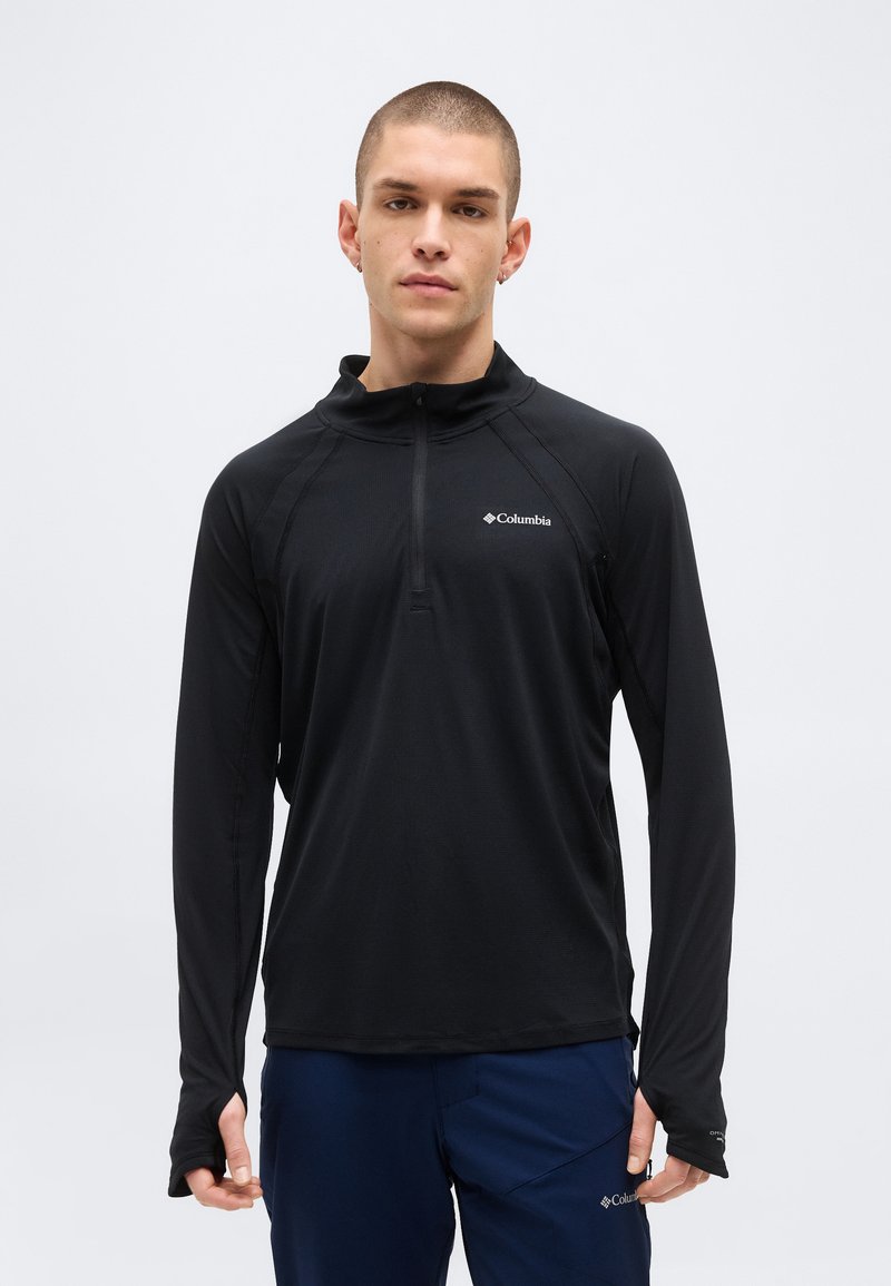 Νέος άνδρας με κοντά μαλλιά, φοράει μαύρο πουκάμισο Columbia με φερμουάρ στο λαιμό και μακριά μανίκια και μπλε σκούρο παντελόνι, στέκεται μπροστά σε απλό φόντο.