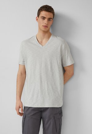 Junger Mann trägt ein hellgraues V-Ausschnitt-T-Shirt und dunkelgraue Cargo-Shorts, steht vor einer einfachen hellgrauen Wand.