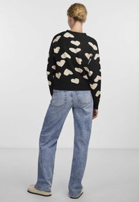 Pull noir avec des motifs de cœurs beige, col rond, poignets côtelés, associé à un jean large bleu clair et des chaussures beige à enfiler.