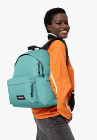 Turkos ryggsäck med en framficka, svarta dragkedjor och justerbara remmar. Bärs över en orange fleece-tröja mot en enfärgad bakgrund.