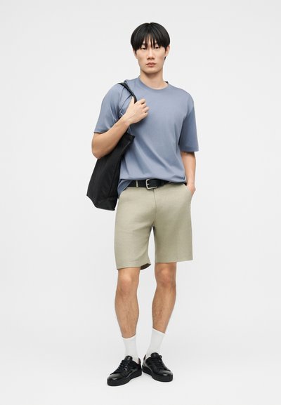 Νέος άνδρας που φορά μπλε μπλουζάκι, μπεζ σορτς, λευκές κάλτσες, μαύρα αθλητικά παπούτσια και κρατά μια μαύρη τσάντα ώμου σε απλό φόντο.