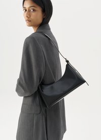 Borsa a tracolla in pelle nera con una forma slanciata e raffinata, dotata di chiusura con zip e di una tracolla sottile, indossata sopra un blazer grigio.