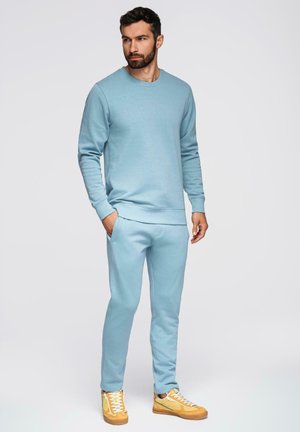 Mężczyzna ubrany w jasnoniebieską bluzę i dopasowane dresy oraz żółte sneakersy, stoi z jedną ręką w kieszeni, patrząc w bok.
