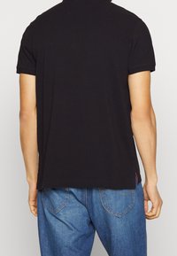 Schwarzes Kurzarm-Polohemd aus Baumwolle mit geradem Saum, dezenten Stickdetails und einem kleinen roten Akzent nahe dem Saum.