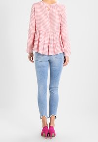 Donna che indossa una blouse a maniche lunghe a scacchi rosa, jeans skinny azzurri con orli sfrangiati e scarpe con tacco alto di un vivido rosa, in piedi di spalle.