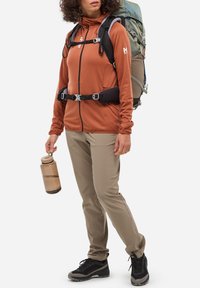 Veste zippée orange avec capuche, pantalon beige, chaussures de randonnée noires et un sac à dos. Tenant une bouteille d'eau brune. Design fonctionnel pour l'extérieur.