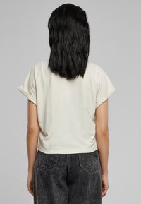 T-shirt crème recadrée avec manches retroussées. Tissu doux, coupe ample. Vue de dos montrant une légère texture, se porte bien avec un jean taille haute foncé.