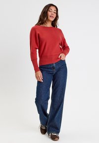 Suéter color óxido con escote amplio y puños acanalados, combinado con jeans de mezclilla azul de tiro alto y zapatillas casuales marrones.