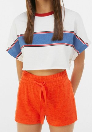 Młoda kobieta w białym krótkim T-shircie z niebieskimi i czerwonymi paskami oraz jasnopomarańczowych krótkich spodenkach ze sznurkiem, stoi z rękami za plecami.